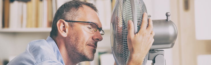 Ein Mann mit Brille sitzt am Schreibtisch und hält sein Gesicht nah vor einen Ventilator zur Abkühlung  – Mehr erfahren zu Abkühlung im Sommer 